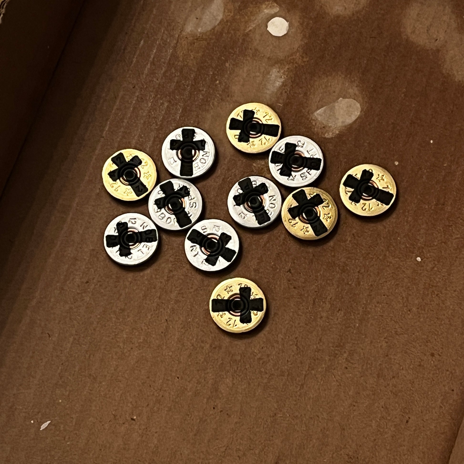 A collection of mixed gold and silver 12-gauge shotgun shell slices scattered on brown cardboard, featuring engraved black cross silhouettes on each authentic thin-cut piece.
