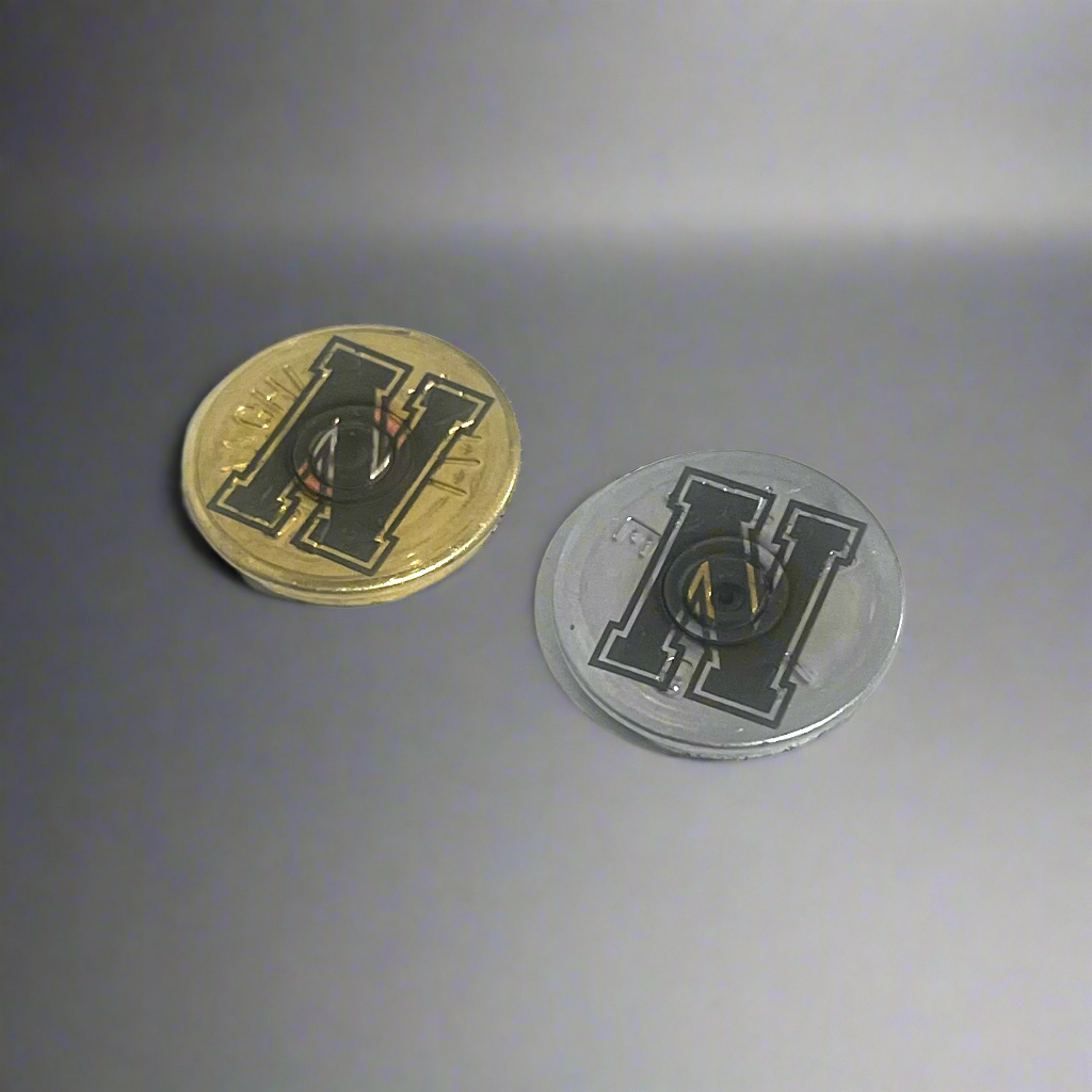 2 round shotgun shell slices, one gold and one silver, engraved with a bold block letter H, angled on a gray background