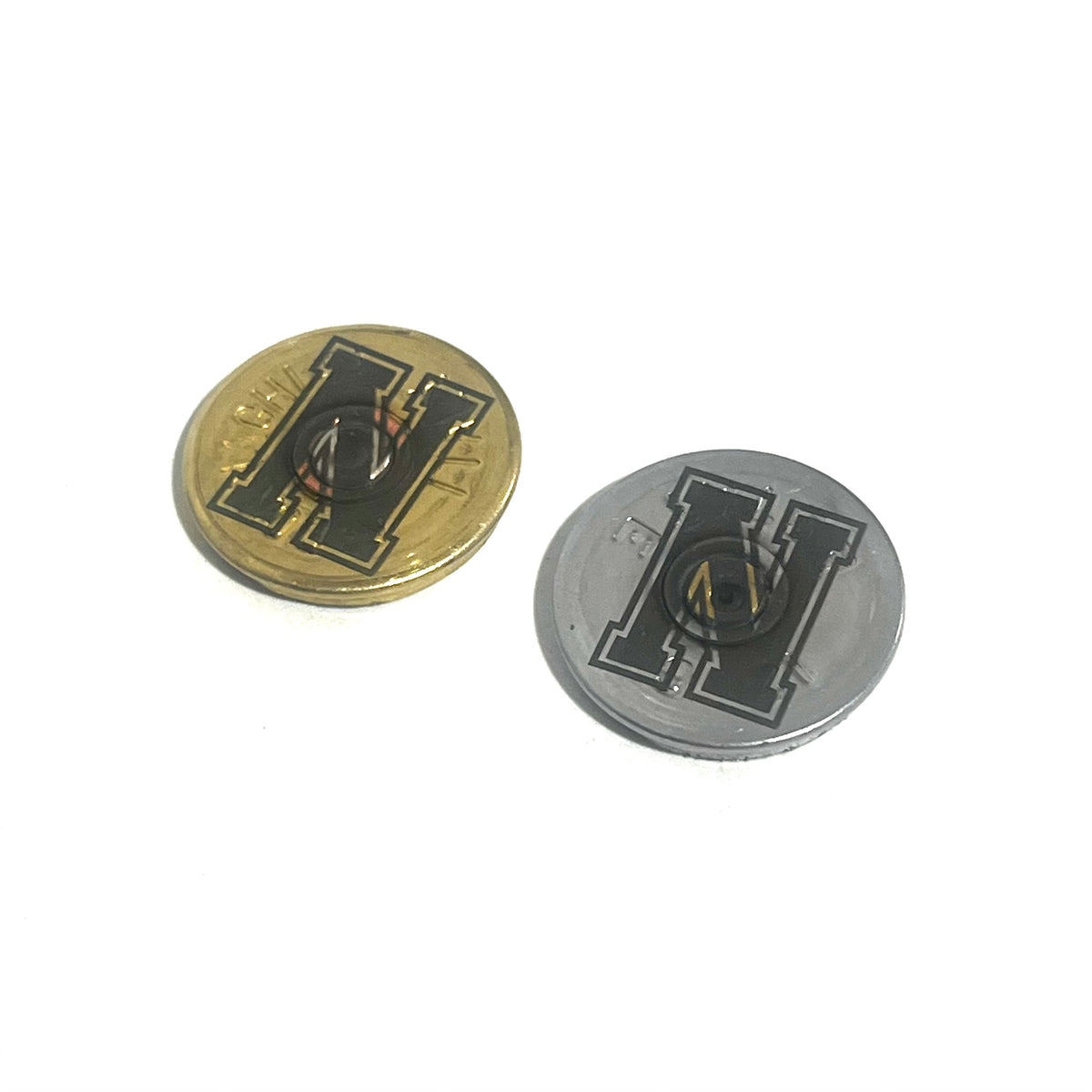 2 round shotgun shell slices, one gold and one silver, engraved with a bold block letter H, angled on a white background
