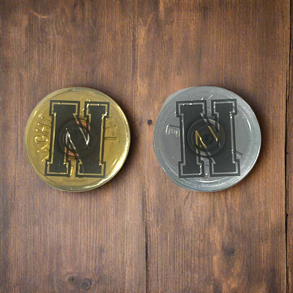 2 round shotgun shell slices, one gold and one silver, each engraved with a bold block letter H, on a wood surface