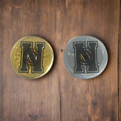 2 round shotgun shell slices, one gold and one silver, each engraved with a bold block letter H, on a wood surface