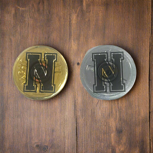 2 round shotgun shell slices, one gold and one silver, each engraved with a bold block letter H, on a wood surface