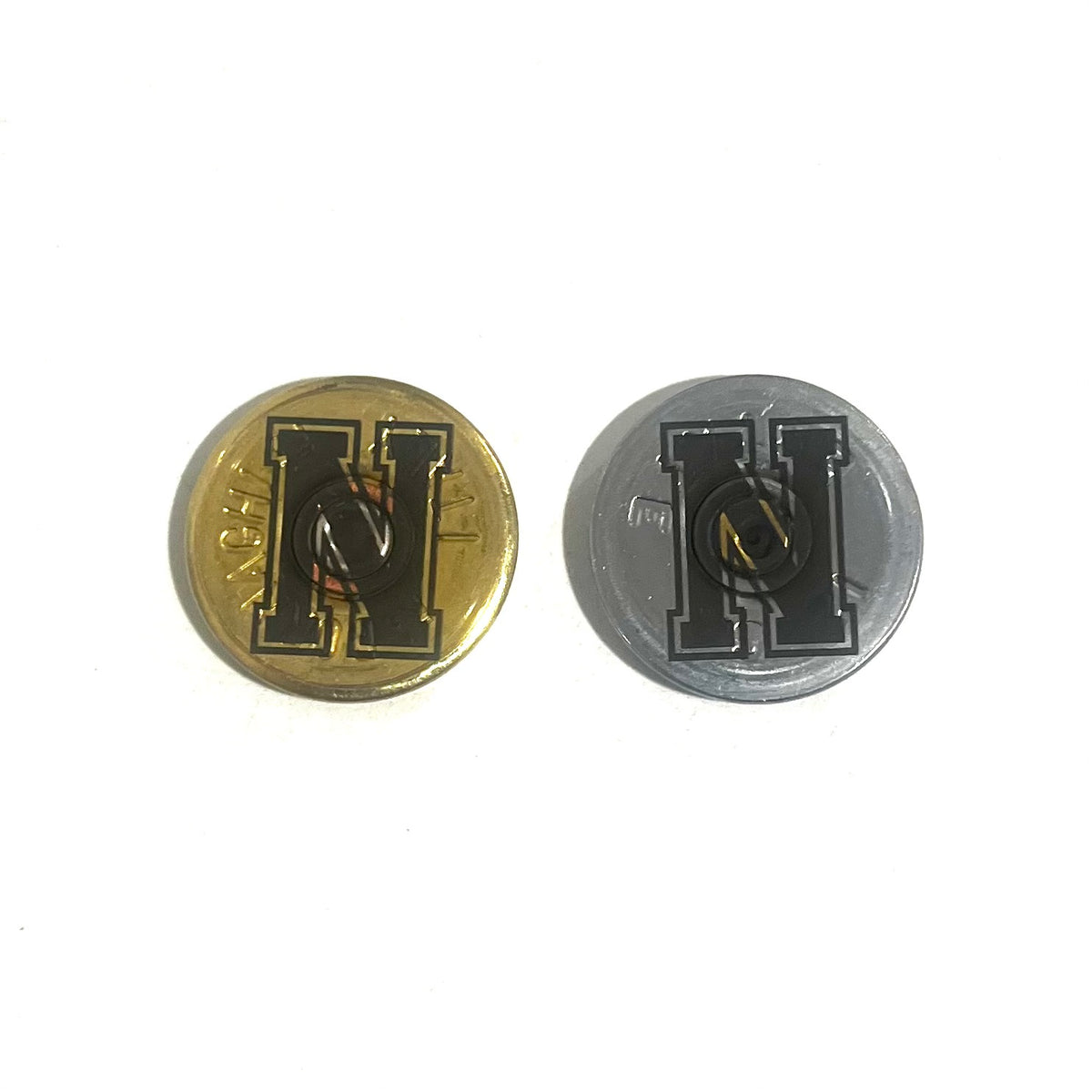 2 round shotgun shell slices, one gold and one silver, engraved with a bold block letter H, on a white background