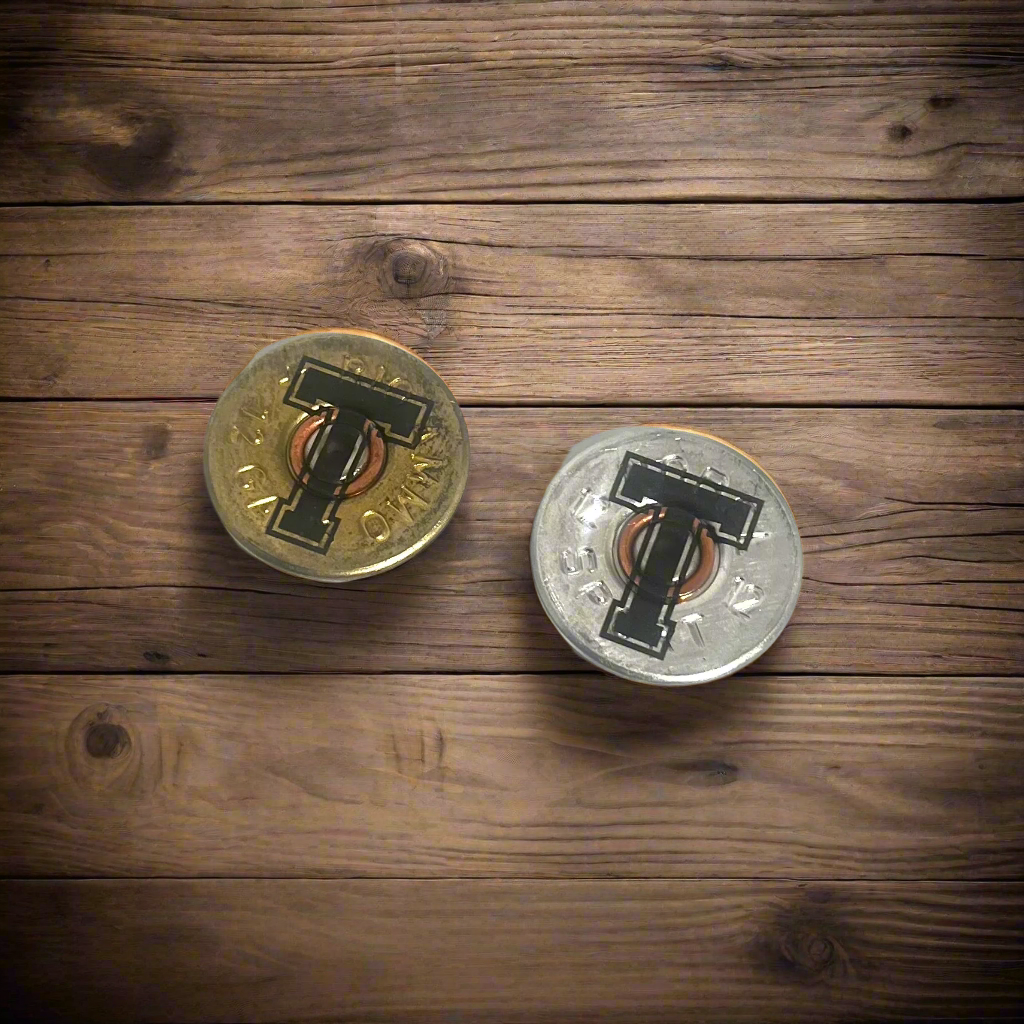 2 round 12 gauge shotgun shell slices engraved with a large letter T, one gold brass and one silver, on a wood surface