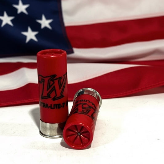 Two red fake shotgun shells positioned in front of a blurred American flag background.
