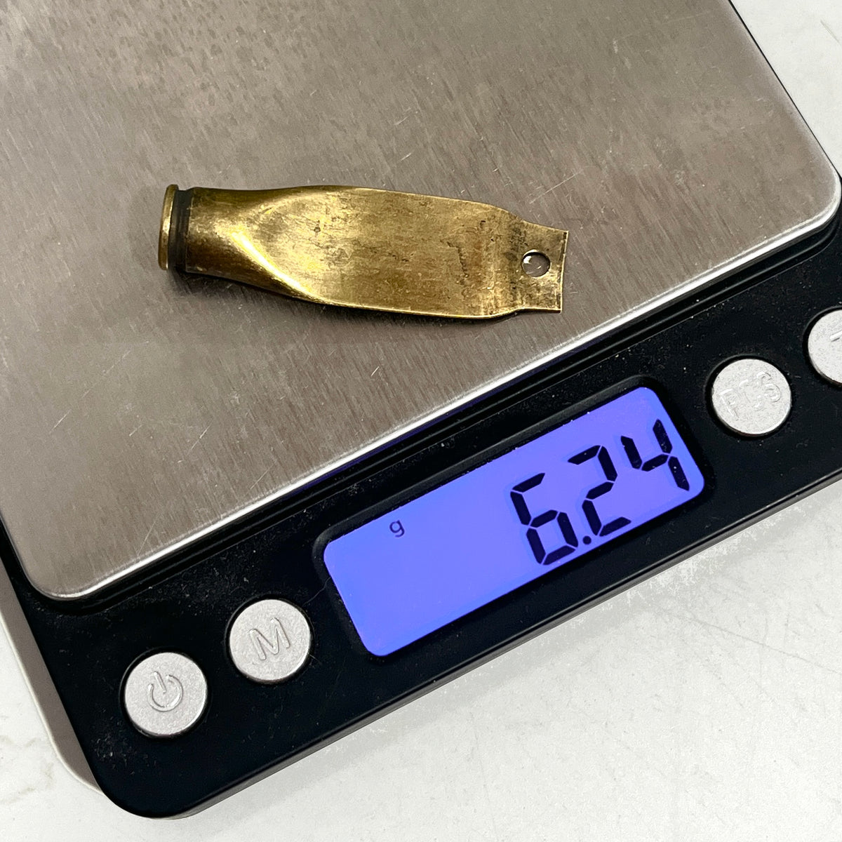 A flattened brass casing with a drilled hole on a digital gram scale showing 6.24 grams