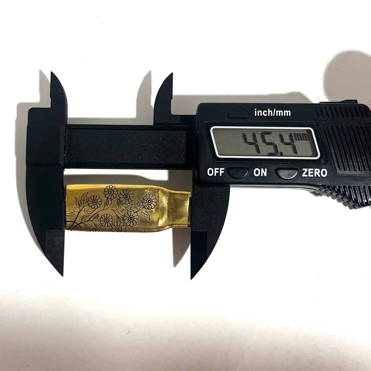 Digital caliper measuring the length of a flattened brass .308 casing engraved with a floral branch design at 45.4 mm