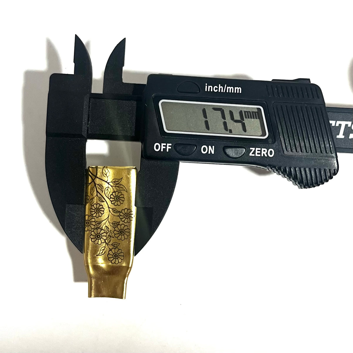 Digital caliper measuring the width of a flattened brass .308 casing engraved with a floral branch design at 17.4 mm