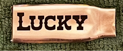 Flattened brass .308 casing engraved with the word LUCKY, laid on a green textured cloth