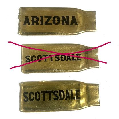 3 flattened brass .308 casings engraved with Arizona and Scottsdale in different font styles, with the middle Scottsdale example crossed out in pink to mark an incorrect font choice