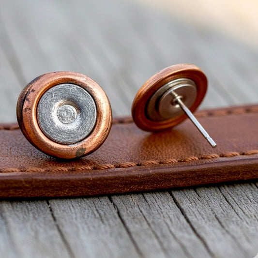 Two copper and silver two-tone spent primers styled as stud earrings resting on a leather surface.