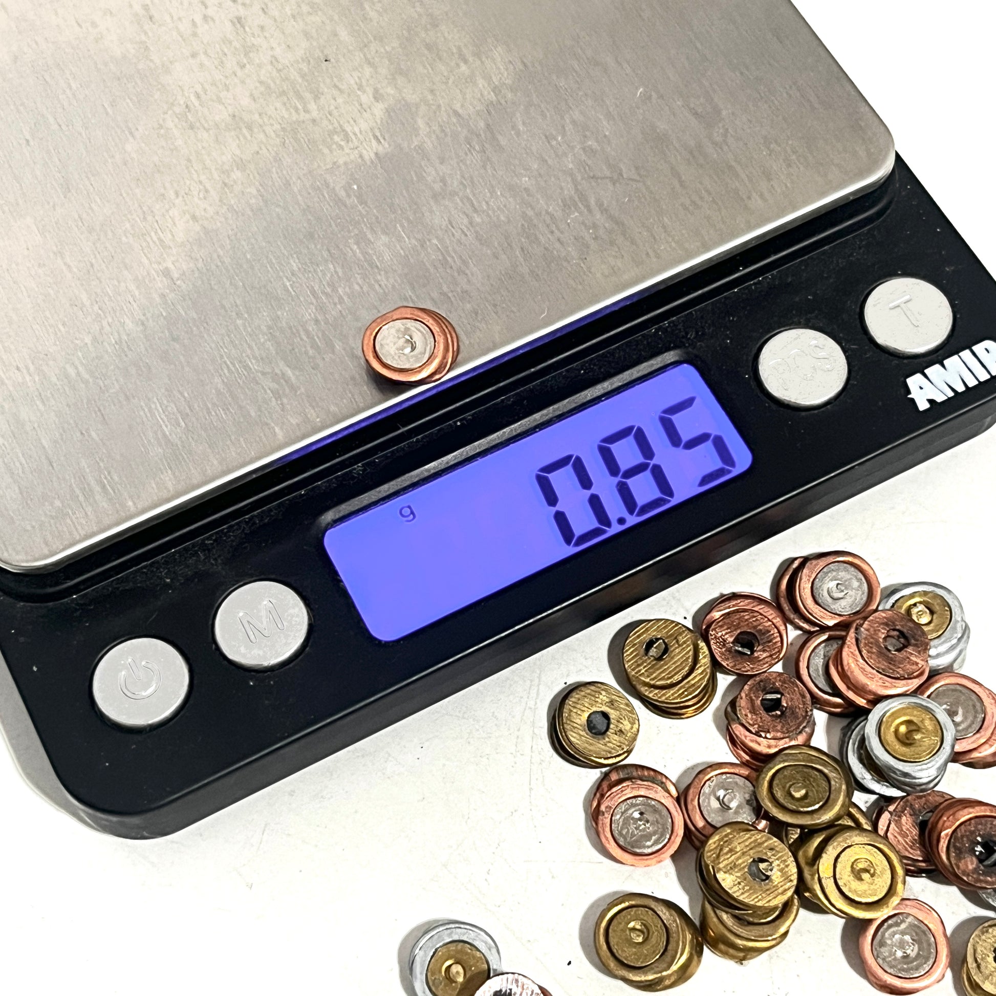 Digital scale measuring the weight of a single spent primer at 0.85 grams, with a pile of spent primers nearby on a white background.