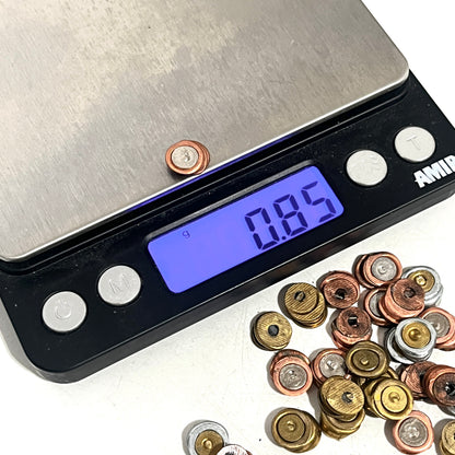 Digital scale measuring the weight of a single spent primer at 0.85 grams, with a pile of spent primers nearby on a white background.