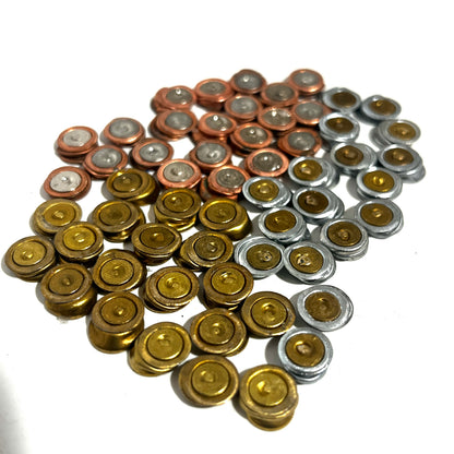 A pile of flattened spent primers in brass, copper, and silver metallic finishes grouped on a clean white backdrop.