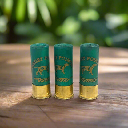 Three green used shotgun casings standing on a wood table with a blurred green foliage background