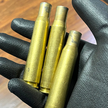 Three uncleaned 50 BMG rifle casings held vertically in a black gloved hand, showing raw tarnish over wood.