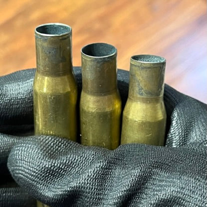 Three uncleaned 50 BMG rifle casings held in a black gloved hand, showing raw tarnish and open case mouths.