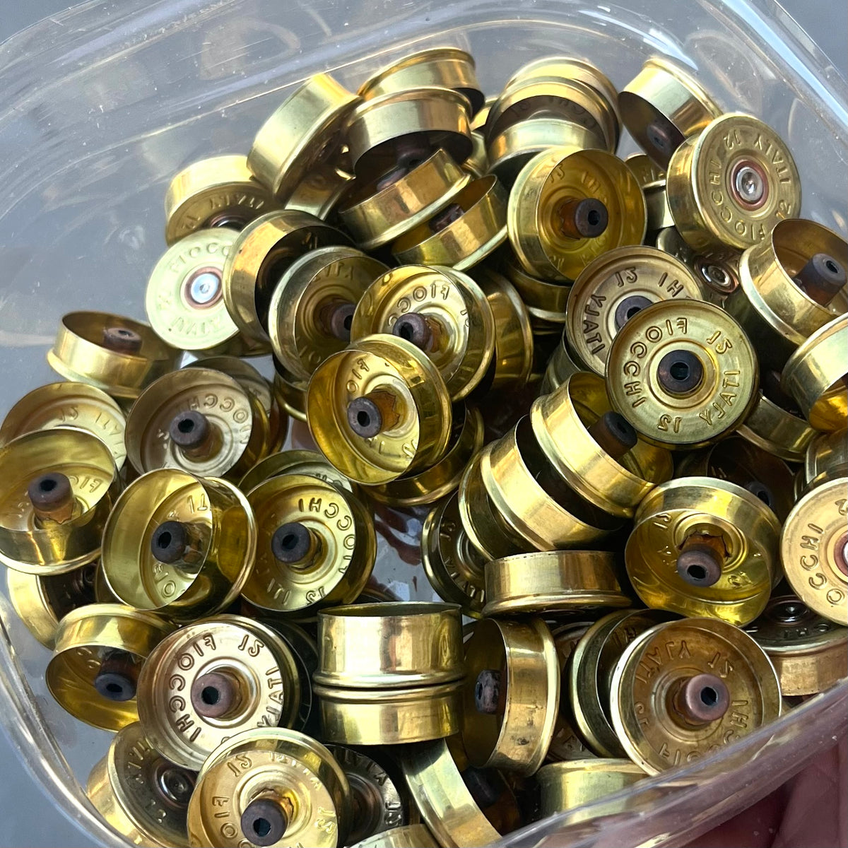Large pile of gold Fiocchi 12 gauge brass end caps with drilled centers in a clear container, one held in a hand