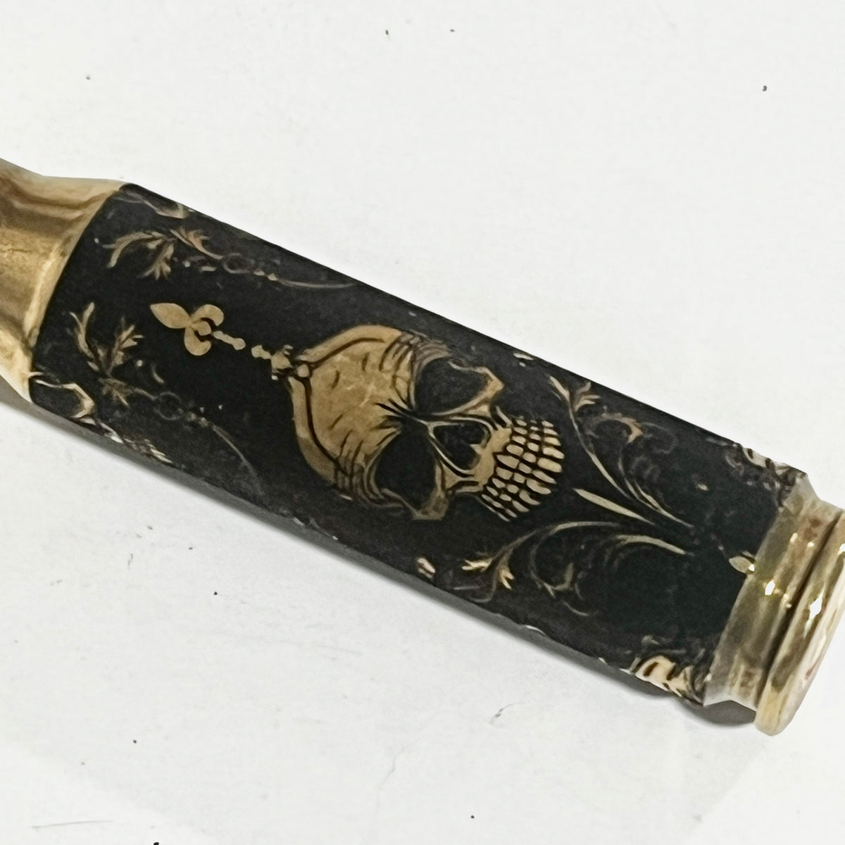 Brass rifle casing engraved with a gold gothic skull and scrollwork design on a black background, angled view