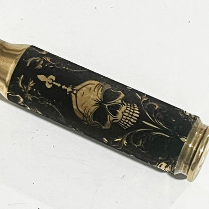 Brass rifle casing engraved with a gold gothic skull and scrollwork design on a black background, angled view