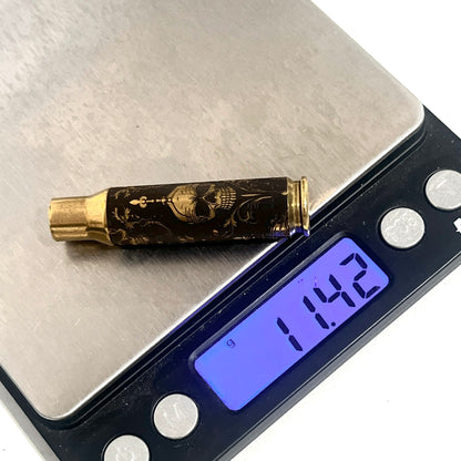 Brass rifle casing engraved with a gold gothic skull design on a digital scale reading 11.42 grams
