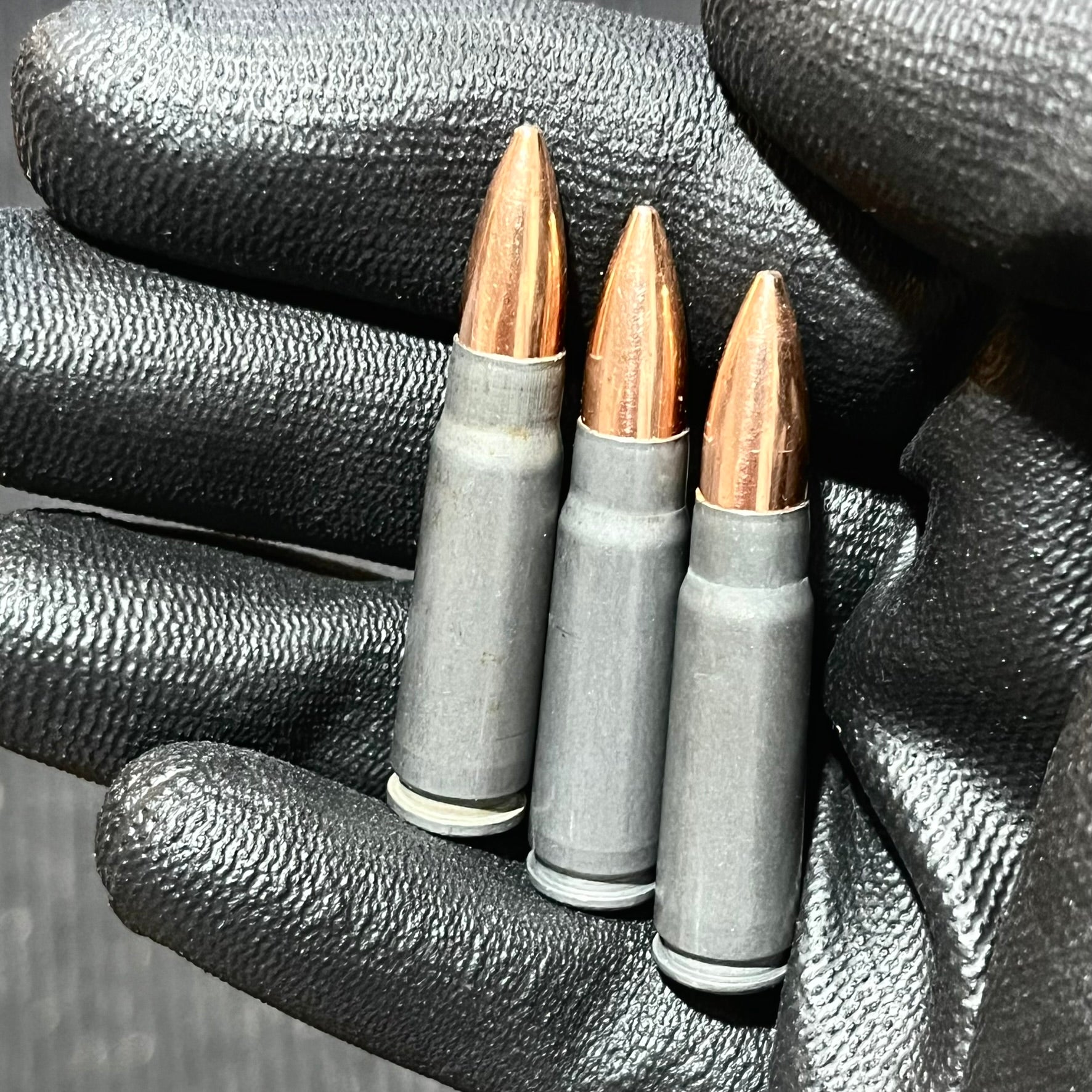 Three gray steel AK47 casings with copper bullets held upright in a black gloved hand.