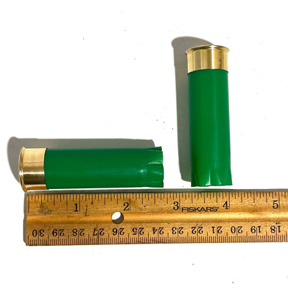2 empty green 12 gauge shotgun shells next to a wooden ruler showing approximately 2.75 inches in length