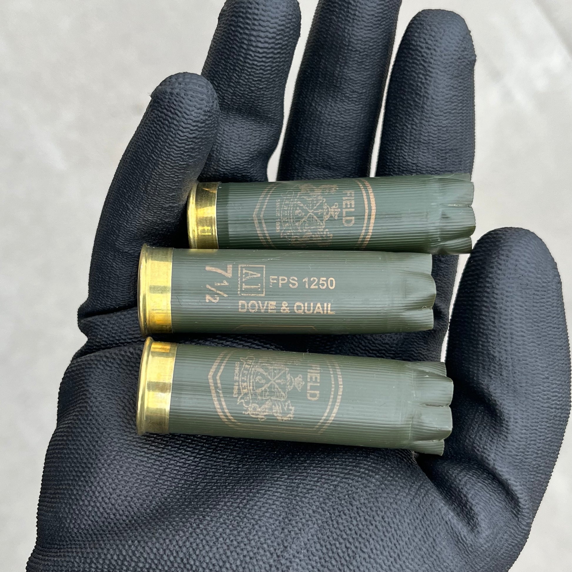 3 empty herters 12 gauge green hulls in black gloved hand showing brass headstamps