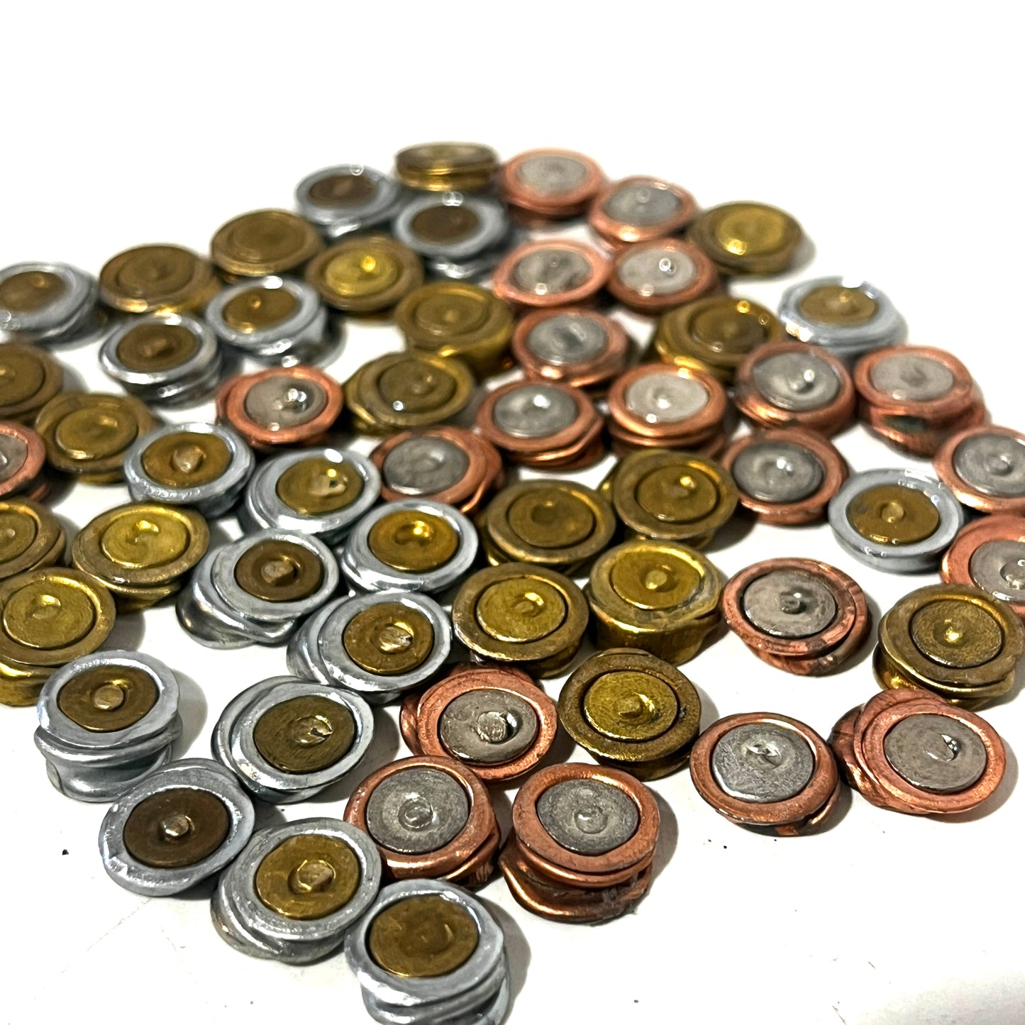A large assortment of inert primer caps in metallic brass, copper, and silver finishes spread over a white background.