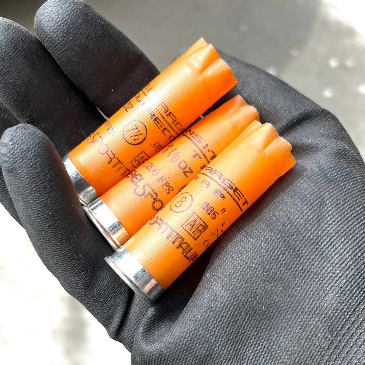 Gloved hand holding three light orange 12 gauge empty shotgun shells printed with Target Recoil and Nobelsport Italia markings