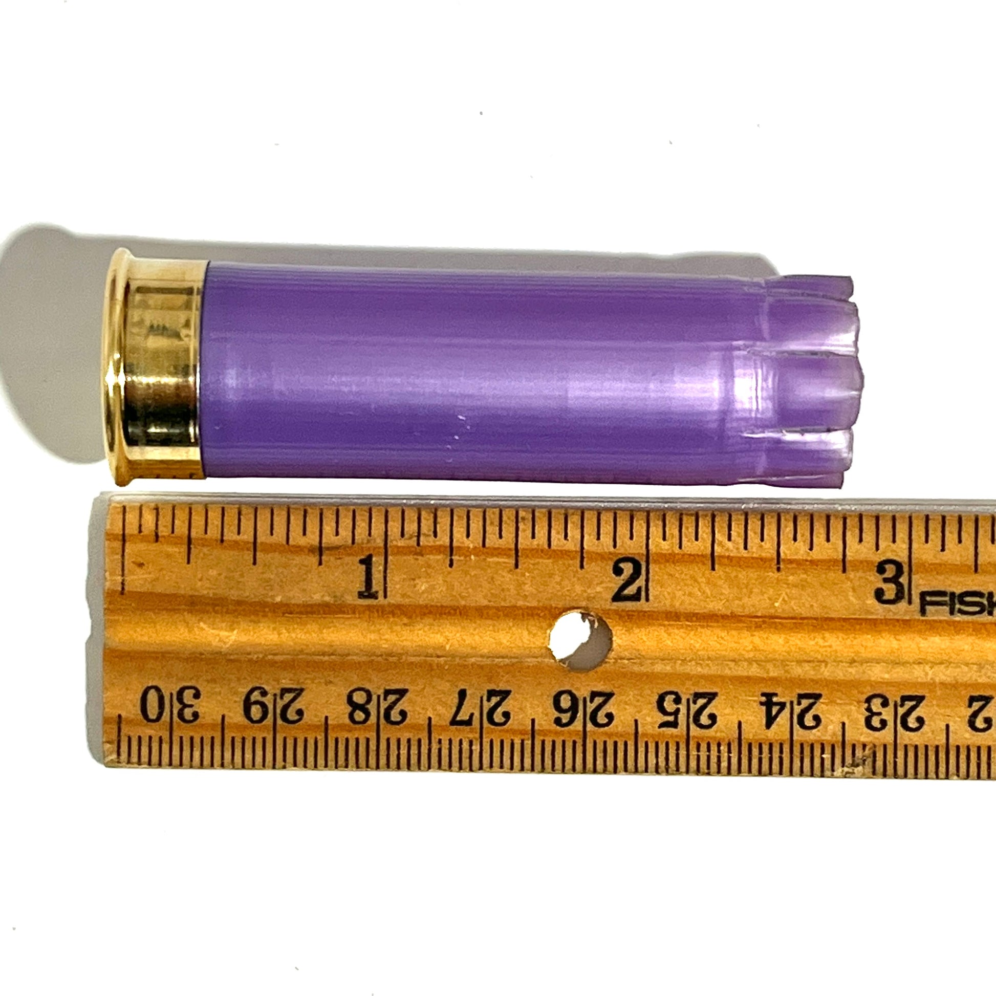 A light purple empty 12ga hull unmarked lying on a wooden ruler measuring about 2 3/4 inches long