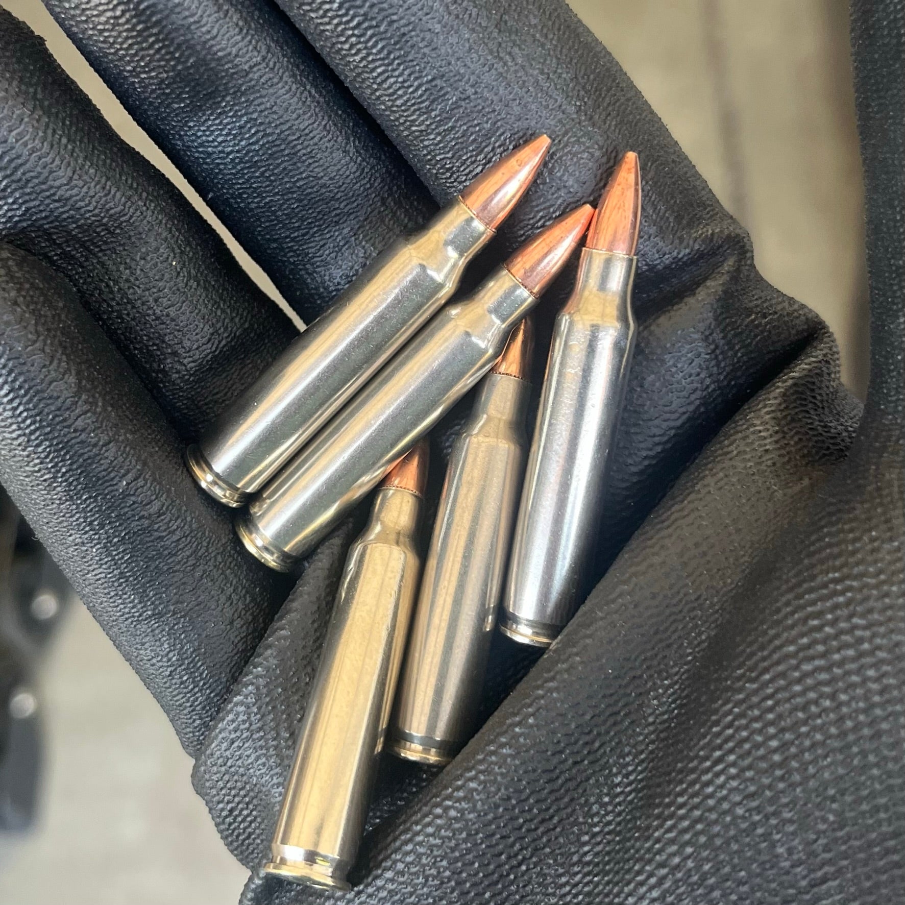 Five mixed nickel and brass .223 dummy rifle cartridges held diagonally in a black gloved hand for film props.