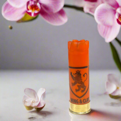 Orange Monarch Wing & Clay 12 gauge shotgun shell with a lion crest logo, standing among pink orchid flowers