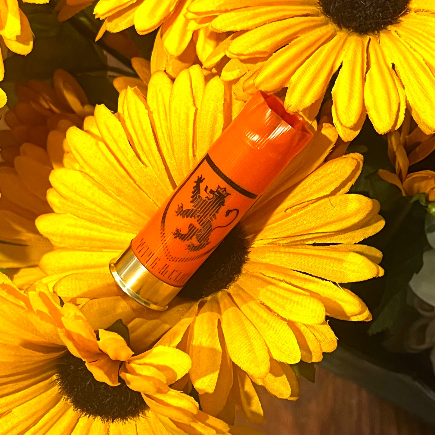 Orange Monarch Wing & Clay 12 gauge shotgun shell with a lion crest logo, nestled among yellow daisy flowers