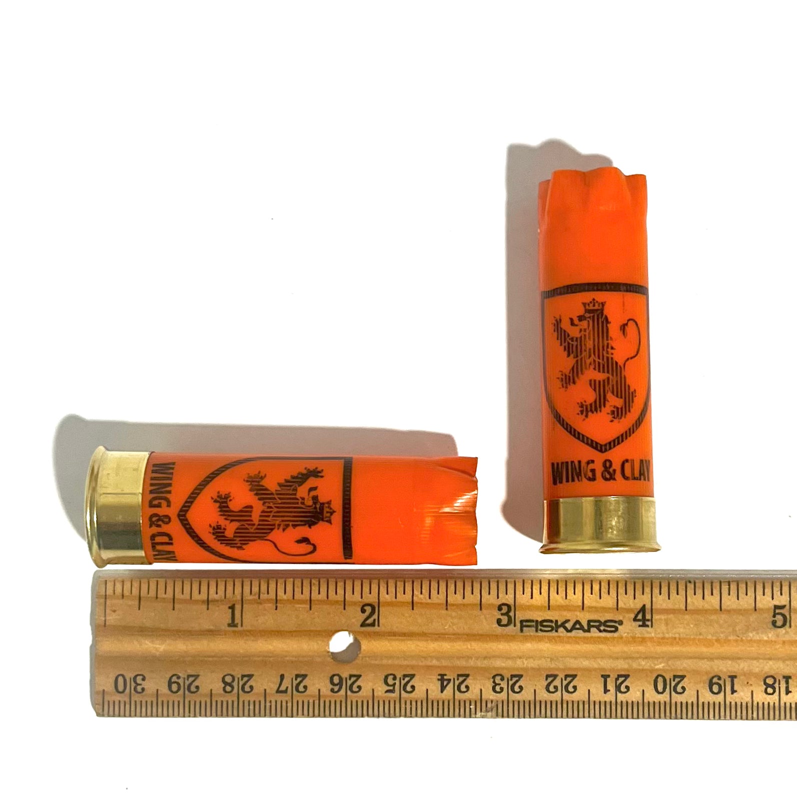 2 orange Monarch Wing & Clay 12 gauge shotgun shells measured on a ruler, one standing upright and one lying down, approximately 2.75 inches