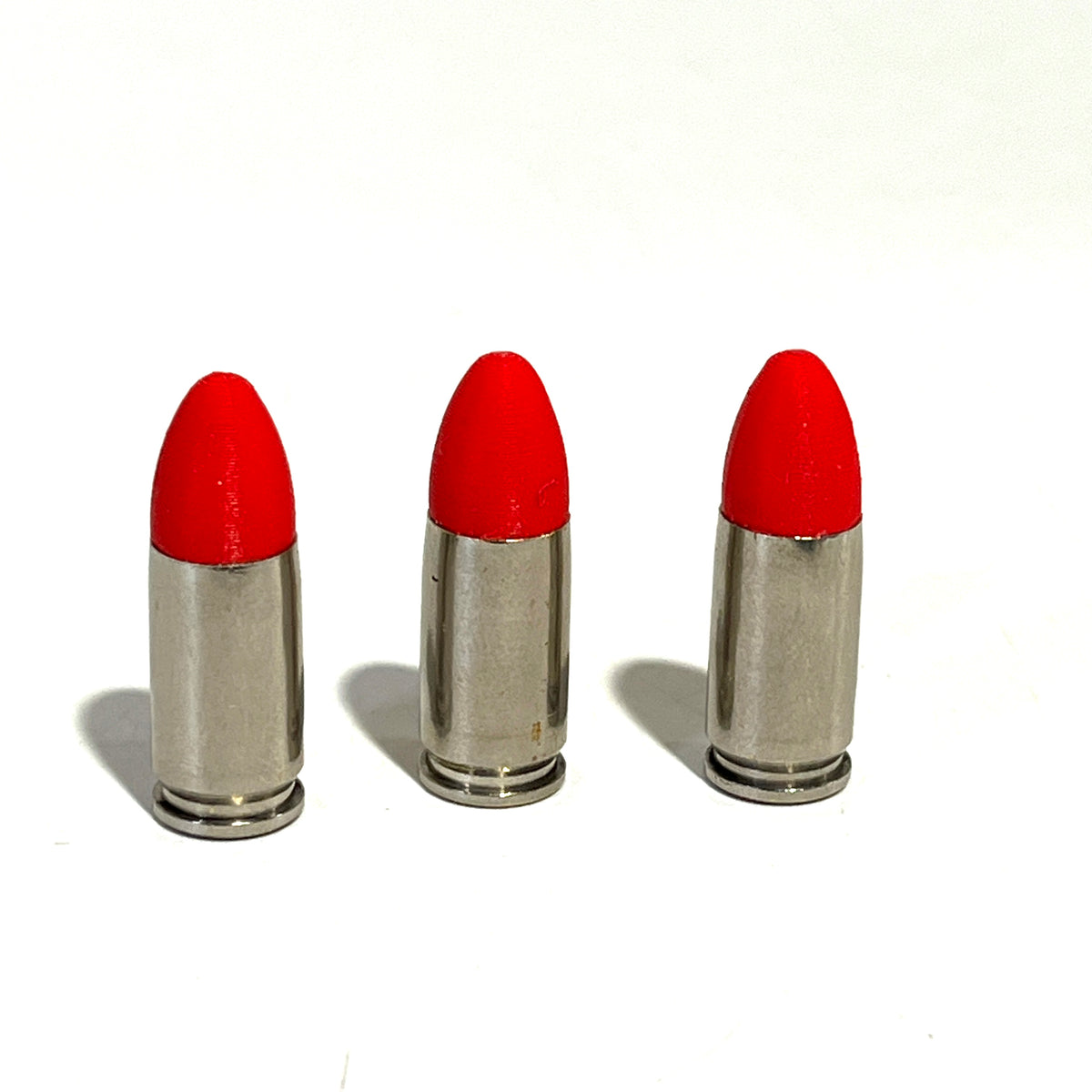 3 fake nickel 9mm Luger dummy rounds with red painted tips standing upright on a white background
