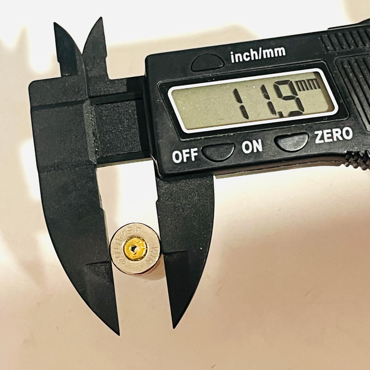 Digital caliper reading 11.9 mm across the rim of a once-fired nickel 45 ACP shell, drilled primer facing up