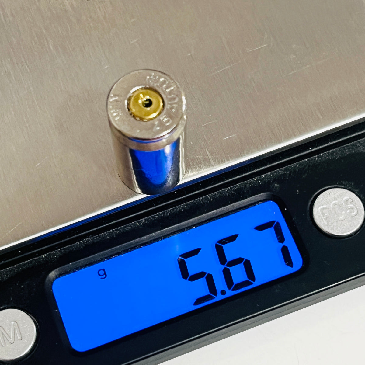 Spent nickel 45 ACP casing with a drilled primer standing on a digital scale reading 5.67 g