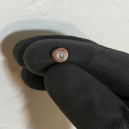 A single round once-fired primers component held by a black gloved hand, showing the copper and silver two-tone details.