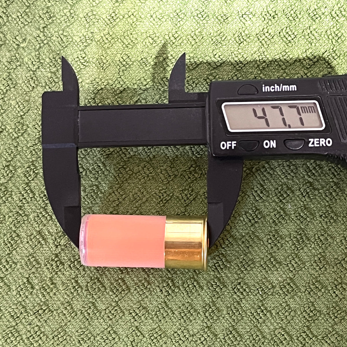 Digital caliper measuring the length of a peach mini shotgun shell at 47.7mm, on green textured towel