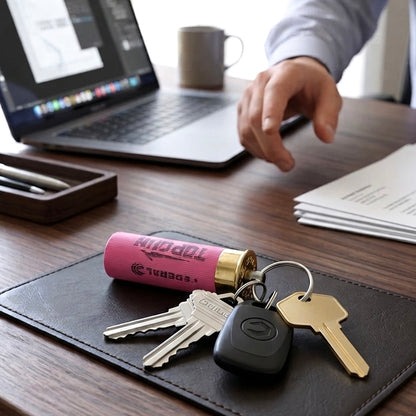 Pink Federal shotgun shell keychain with human hand, keys on desk, laptop, papers and pens.