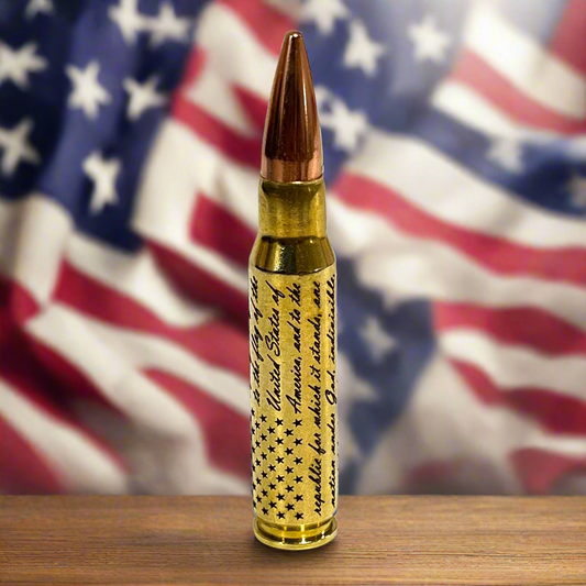 Brass .308 WIN cartridge engraved with the Pledge of Allegiance and a star flag design, standing on a wood table in front of an American flag