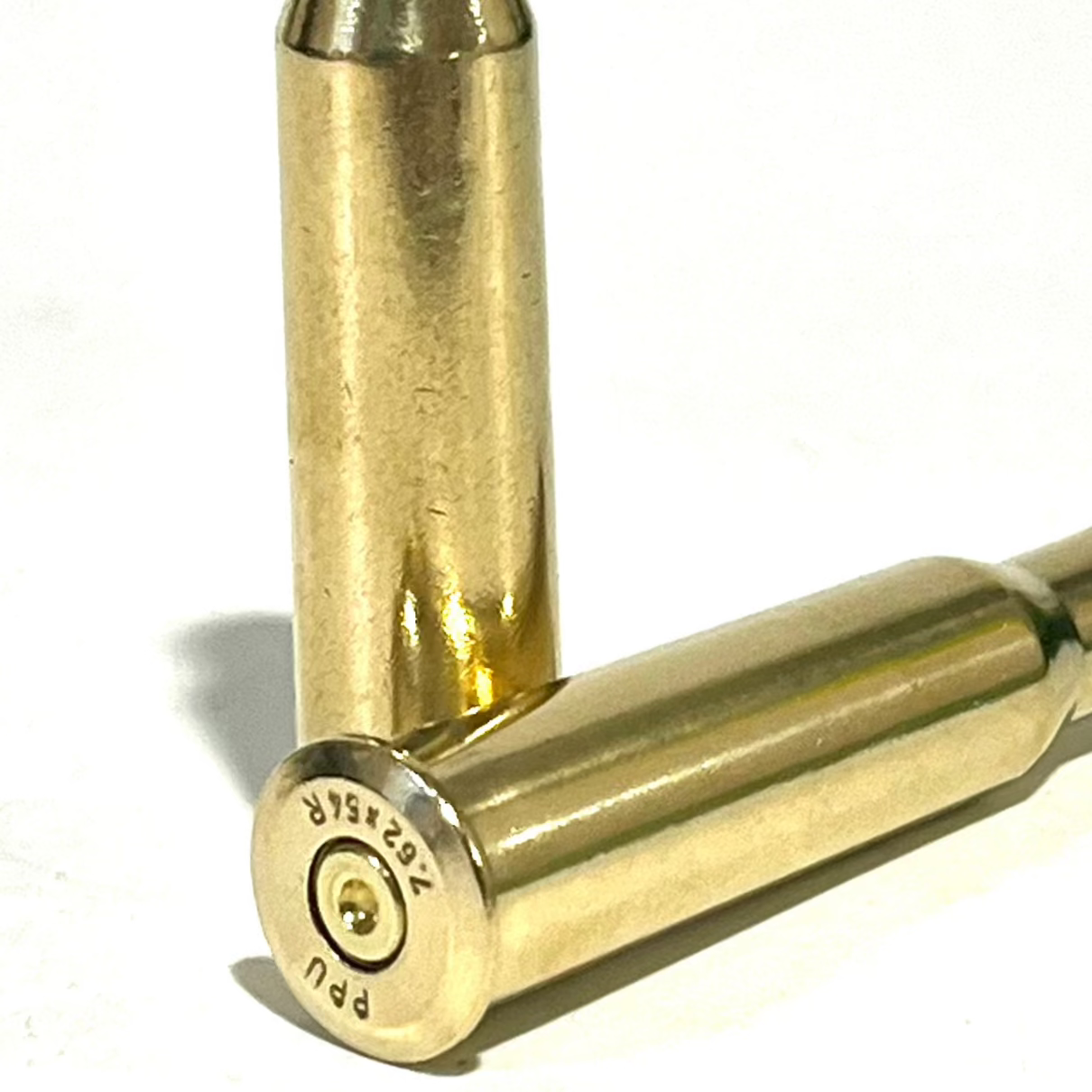 Polished 7.62x54R empty brass casing lying in foreground showing PPU headstamp and cleaned, visibly dimpled, inert primer. White background.
