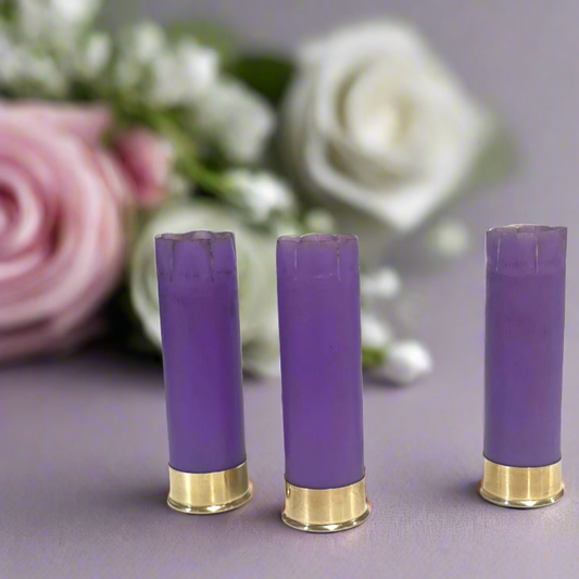 3 purple 16 gauge shotgun shell hulls with brass bases standing in front of blurred pink and white roses