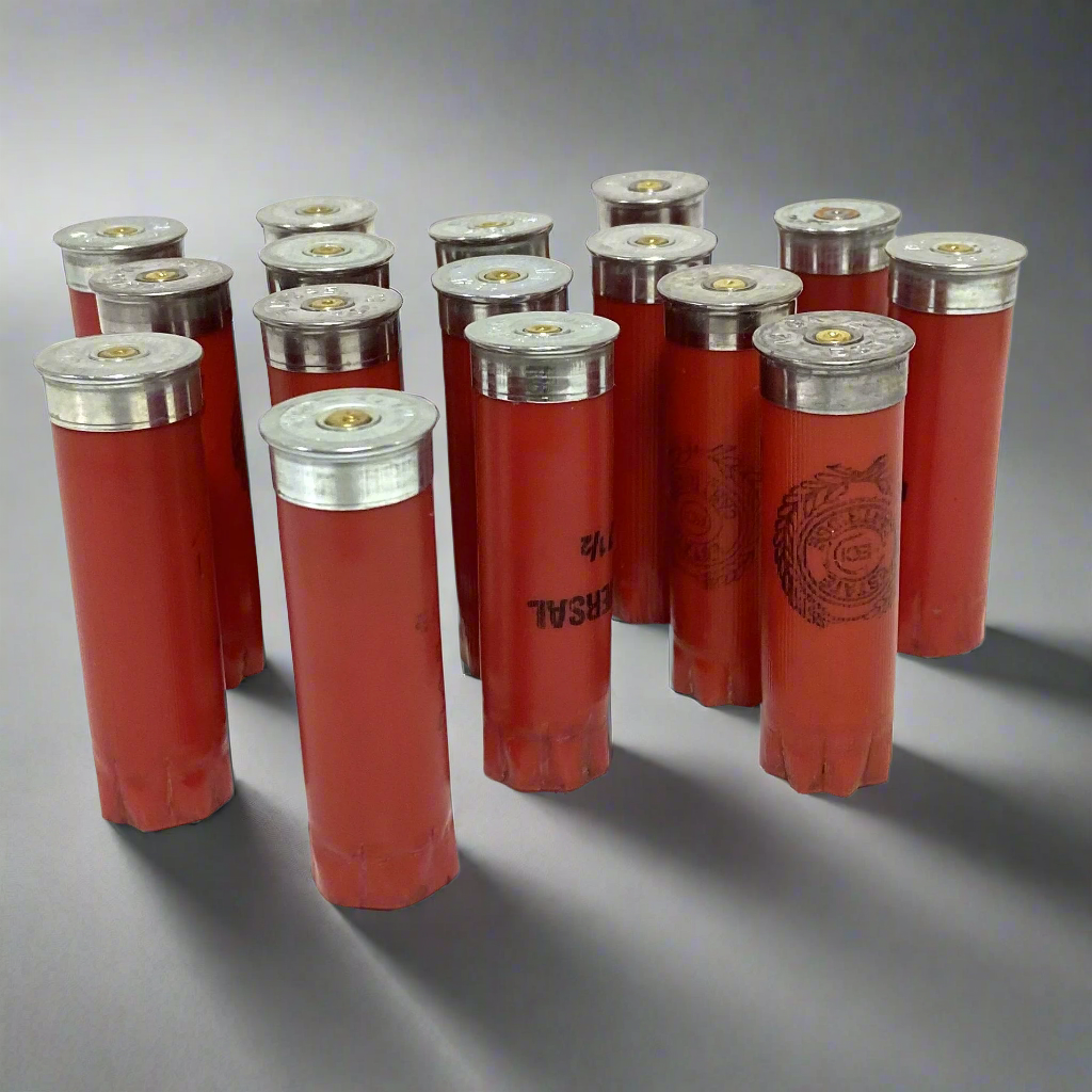 A cluster of twelve recovered, once-fired red 12-gauge empty shotgun shells standing upright on a grey background. The ribbed plastic hulls feature silver-colored bases, star crimps, and black printed graphics, including "UNIVERSAL" text and circular crest logos. Studio lighting casts distinct, long shadows to the right
