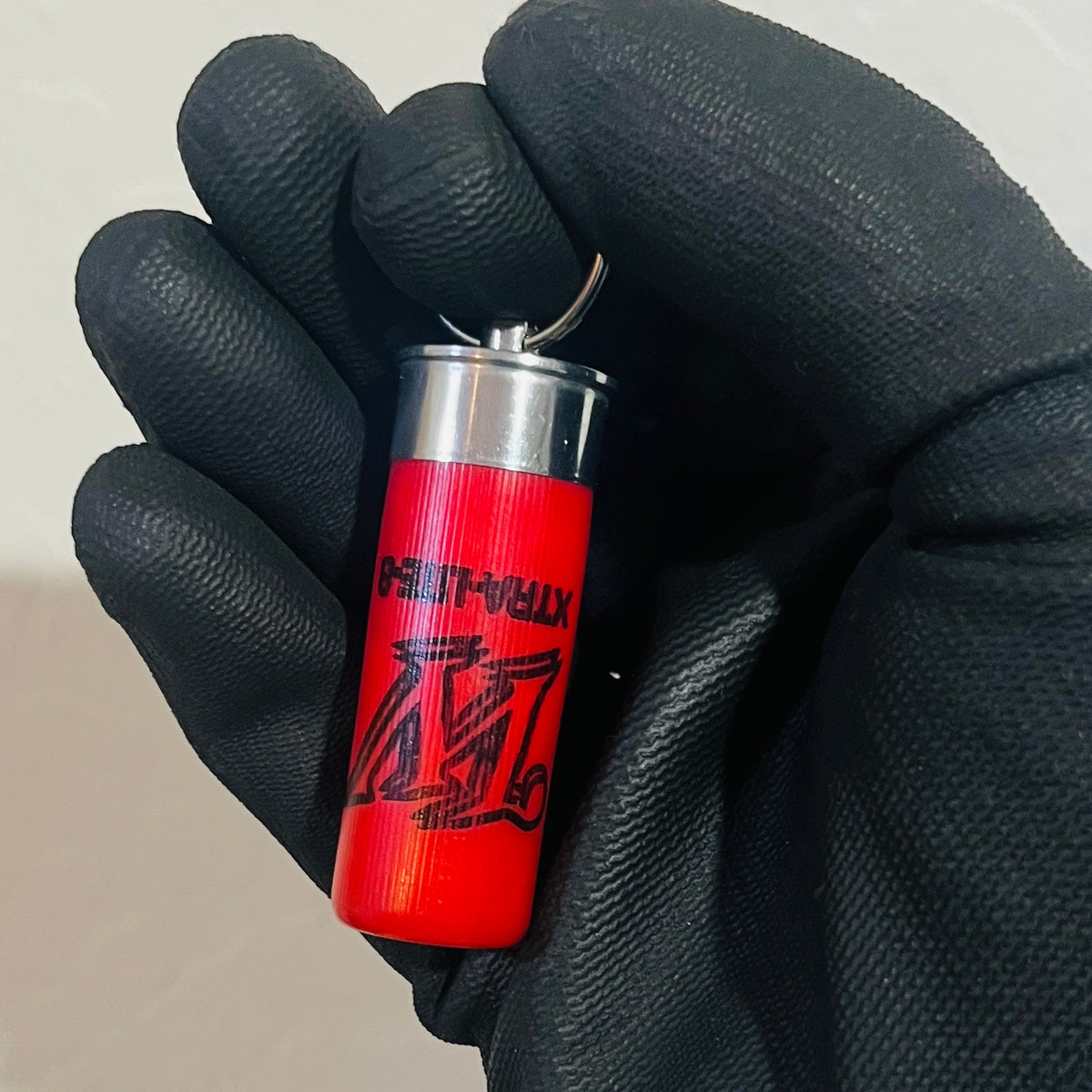 Red empty shotshell keychain held upright in a black textured glove, showing the W logo and silver base
