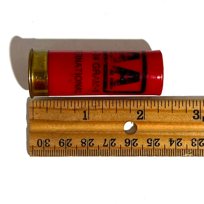Single red Winchester AA International 12 gauge shotgun dummy round laid against a wooden ruler measuring about 2.7 inches