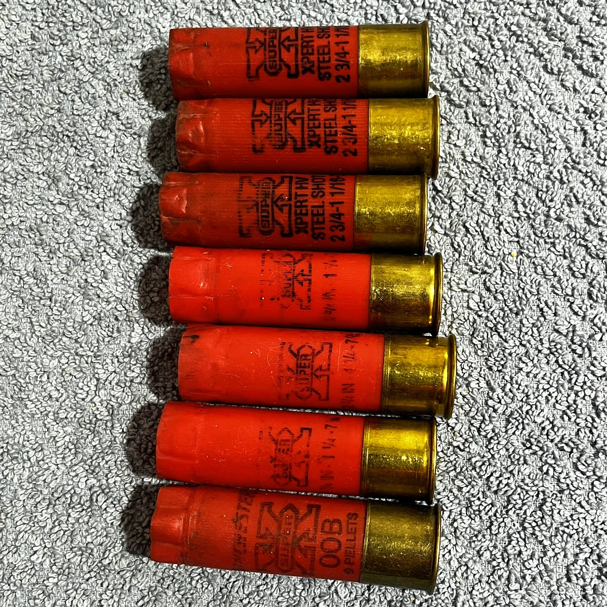 Seven red Winchester Super X 12 gauge empty shotgun shells with gold high brass bases lying on a gray carpet