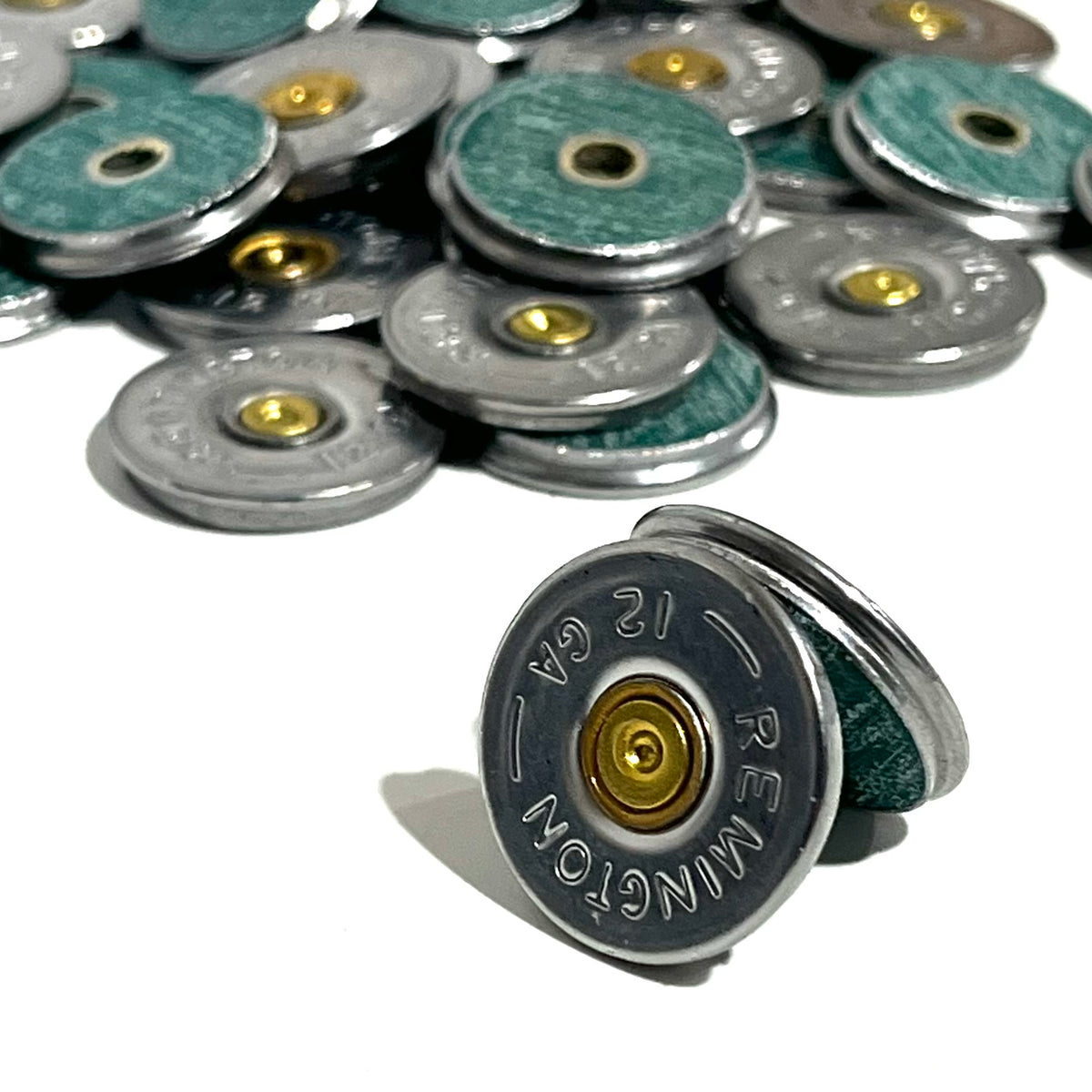 A Remington 12 gauge bullet slice standing on its edge showing the thin green hull material and brass primer, with a pile of additional slices blurred in the background