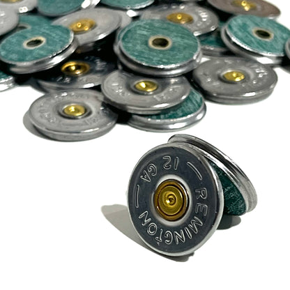 A Remington 12 gauge bullet slice standing on its edge showing the thin green hull material and brass primer, with a pile of additional slices blurred in the background